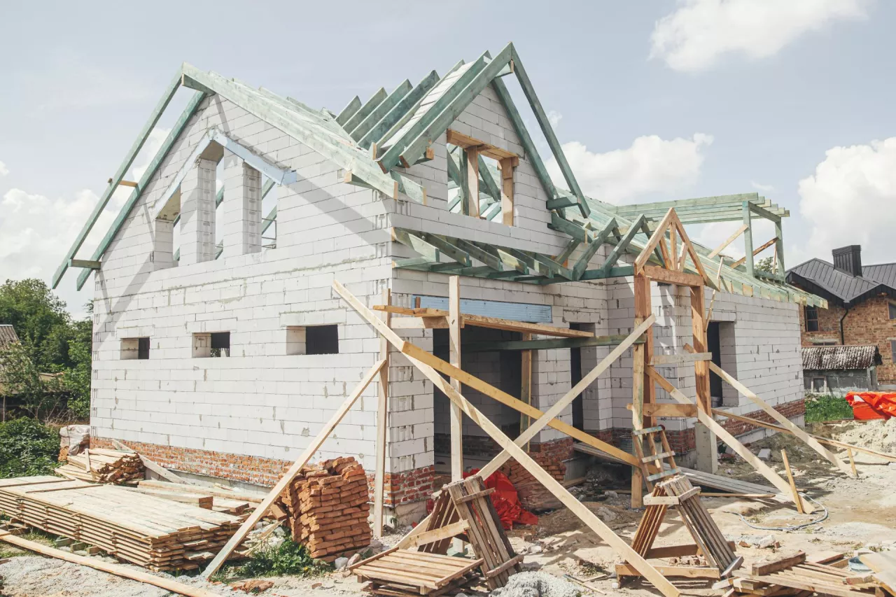 Unfinished modern farmhouse building. Wooden roof framing of mansard with dormer and aerated concrete block walls with windows. Rafters and beams on blocks. New house construction